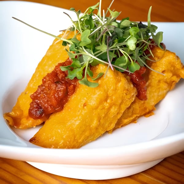 Two golden vegan empanadas topped with red salsa and fresh microgreens on a white plate.