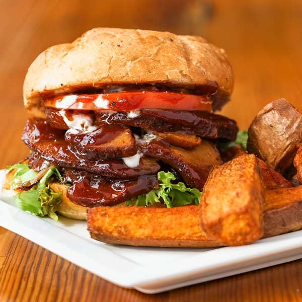 A vegan BBQ pulled pork sandwich with lettuce, tomato, and creamy sauce, served with sweet potato fries.